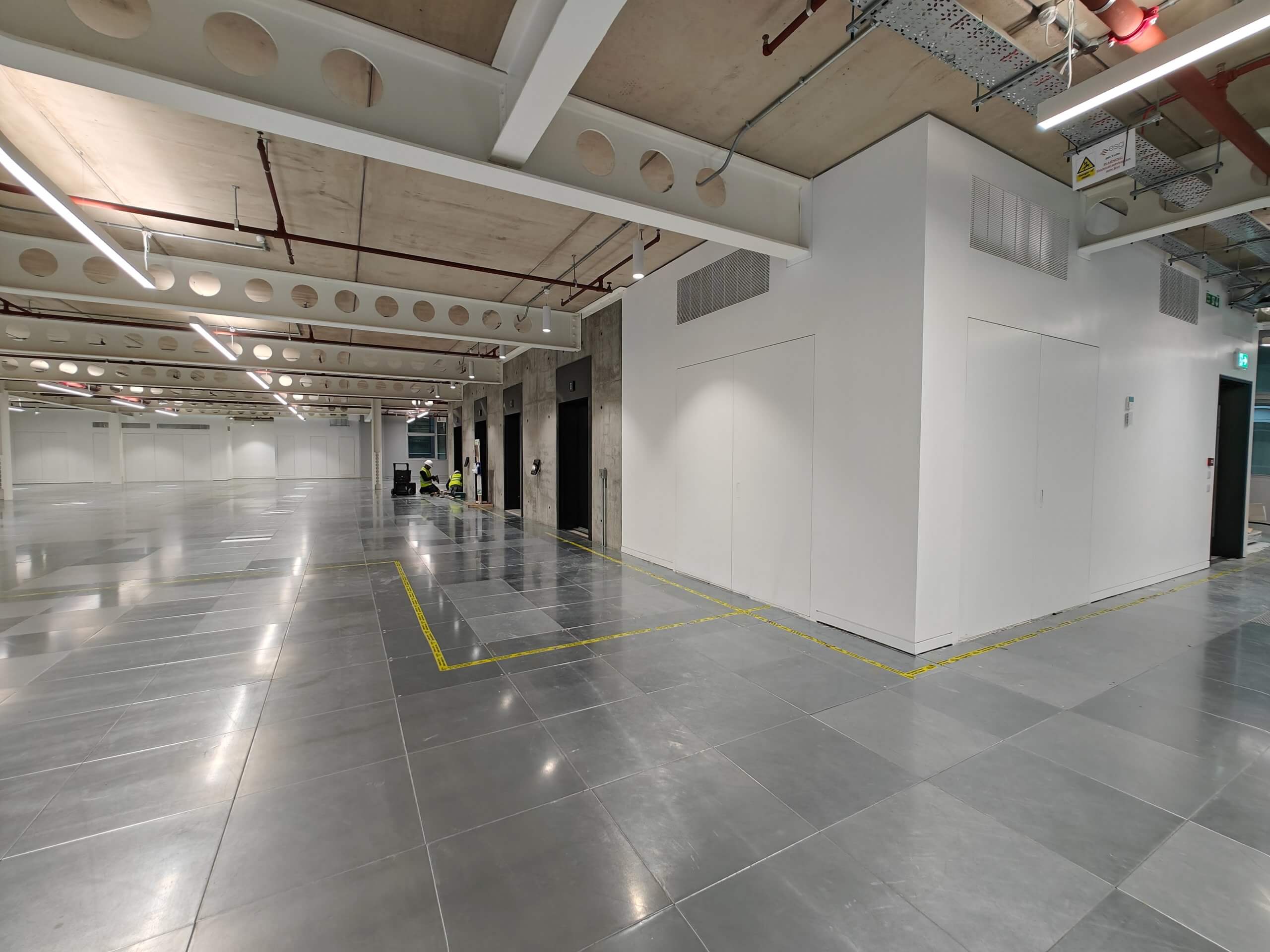 Modern commercial space with Christo Riser Doors installed in the foreground and background. Construction workers are seen working near a lift shaft, while exposed ceiling beams and a clean, spacious design complete the space.
