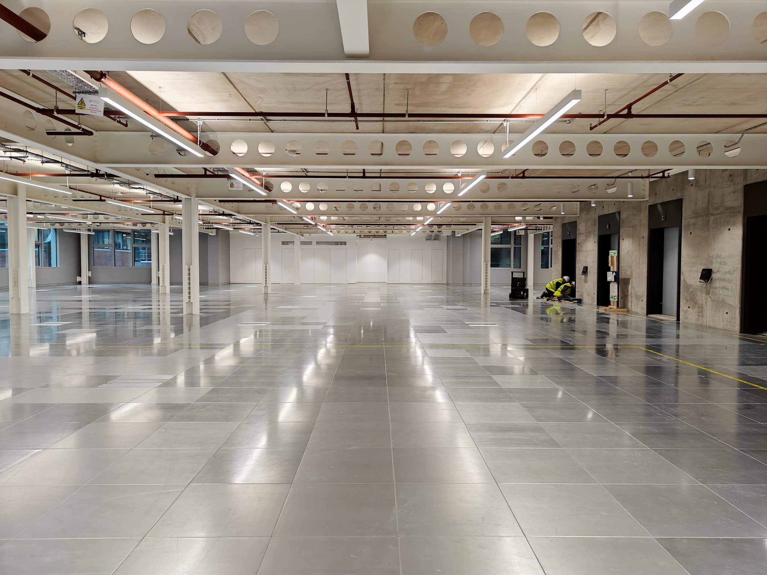 Construction worker in a high-visibility vest and hard hat inspecting an open Christo Riser Door in a modern commercial space. Additional closed Christo Riser Doors and exposed ceiling infrastructure are visible in the background.