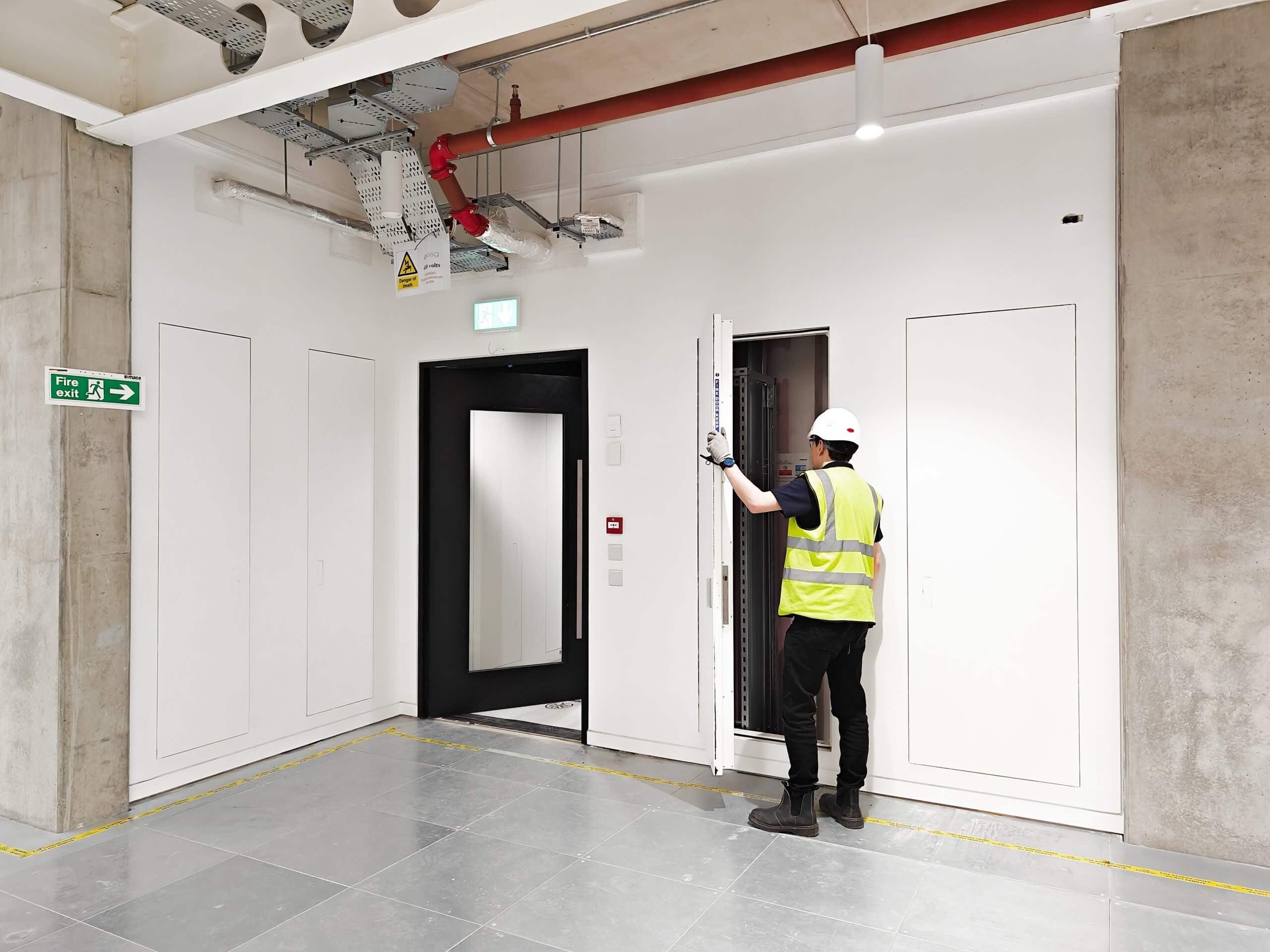 Construction worker in a high-visibility vest and hard hat inspecting an open Christo Riser Door in a modern commercial space. Fire exit signage and exposed ceiling infrastructure are visible, with a clean and professional environment.
