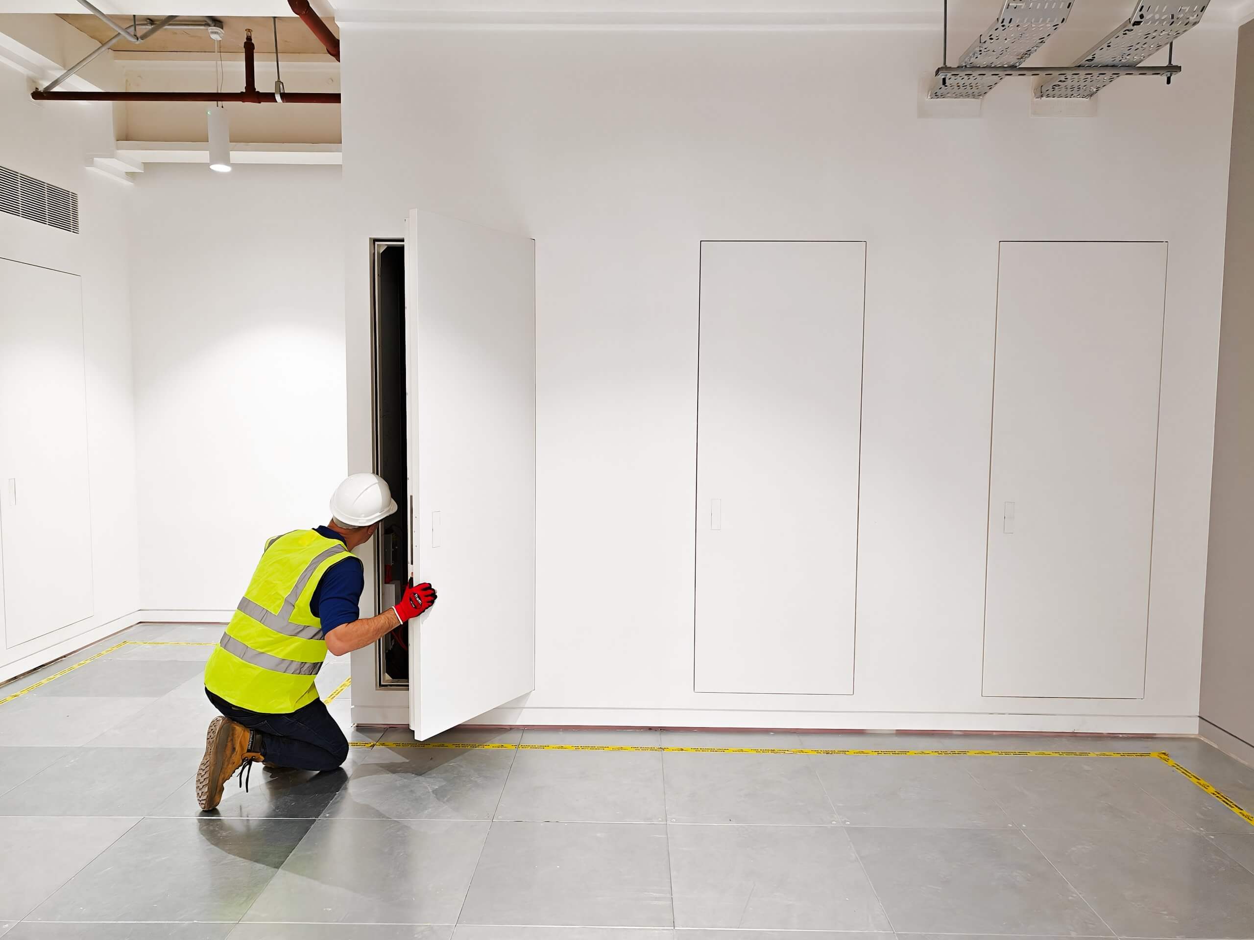 Construction worker in a high-visibility vest and hard hat inspecting an open Christo Riser Door in a modern commercial space. Additional closed Christo Riser Doors and exposed ceiling infrastructure are visible in the background.
